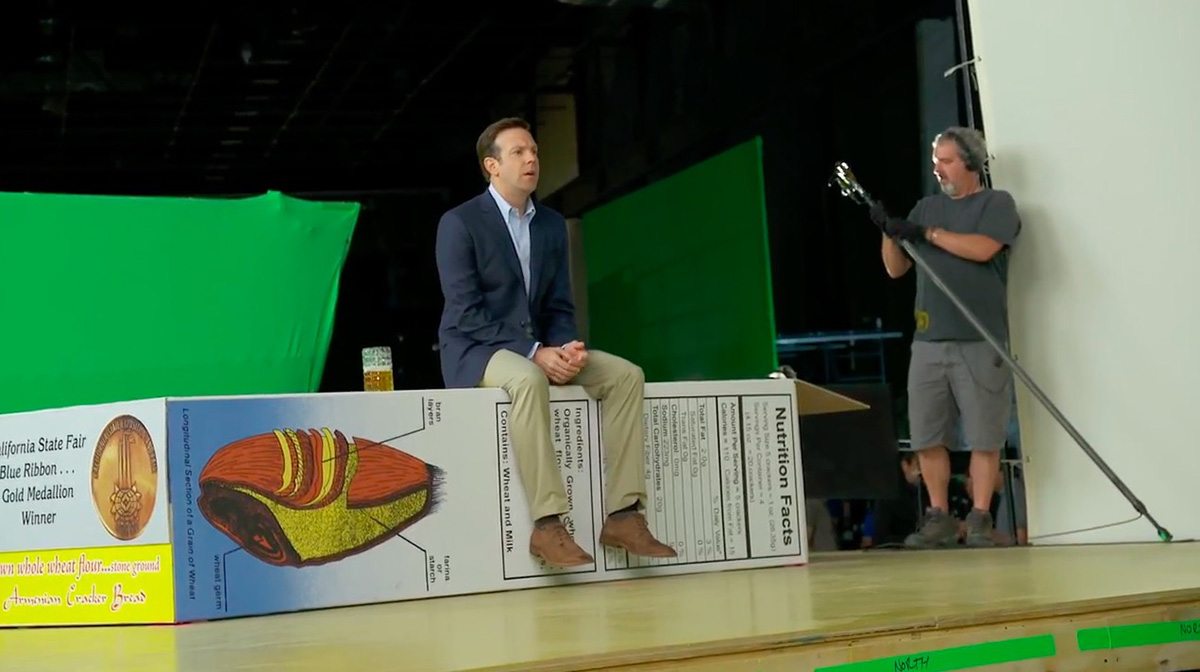 A screenshot of Jason Sudeikis from b-roll footage shows him performing against greenscreen on an oversized set.